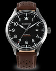 Close-up of a Wancher Flugel Automatic Pilot Watch on a person's wrist, featuring a black dial with white numerals and hands, and a brown Rally Strap leather strap.