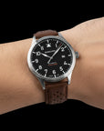 A person's wrist wearing the Wancher Flugel Automatic Watch, a pilot-style timepiece with a contrasting black dial and warm brown leather strap.