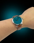 Wrist wearing a watch with a blue face and brown leather strap against a gradient background