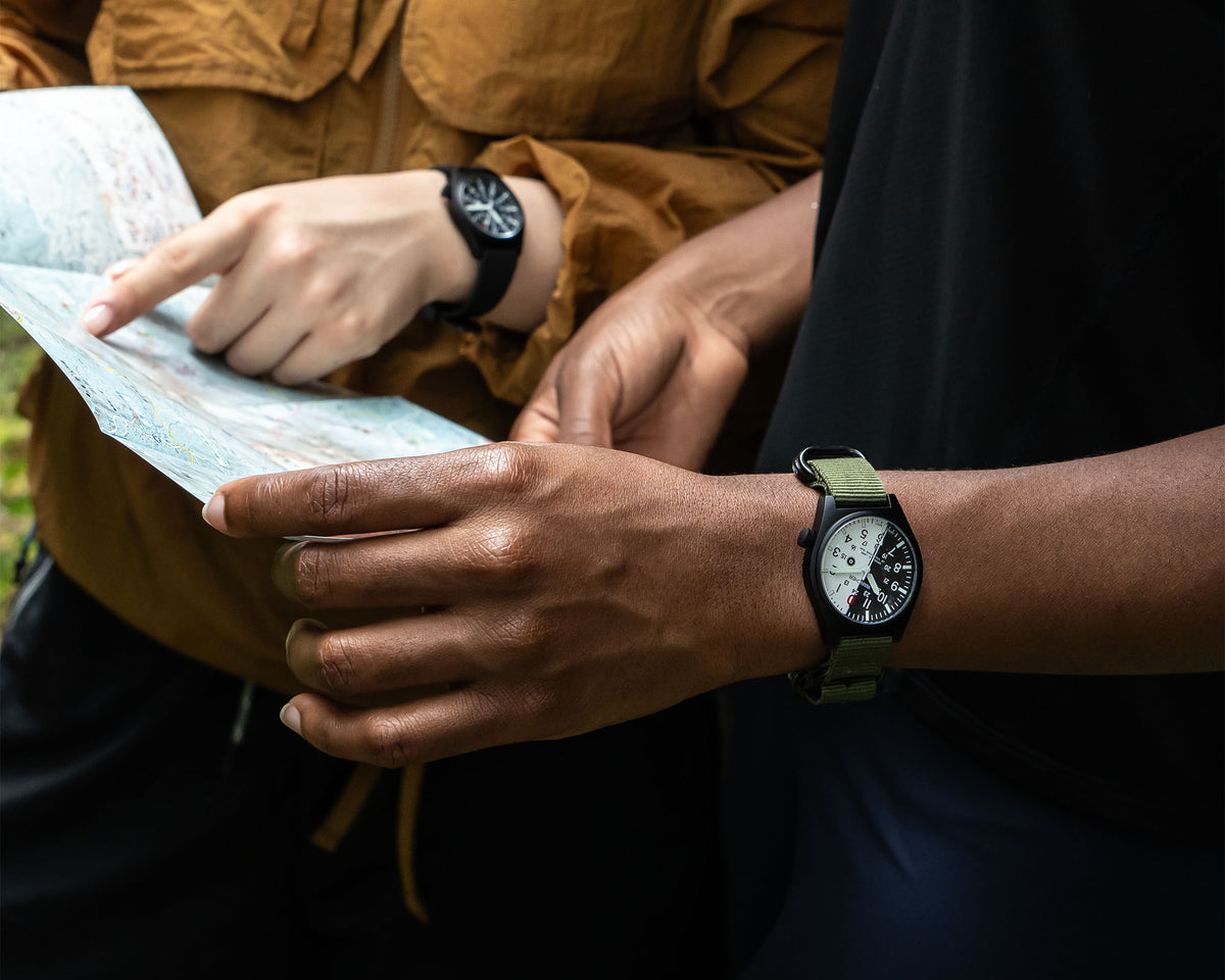 A close-up lifestyle shot of a person wearing an olive green and black outdoor watch while looking at a map with another person in a brown jacket. Navigating with Wancher Gurkha tilted dial hiking watch 