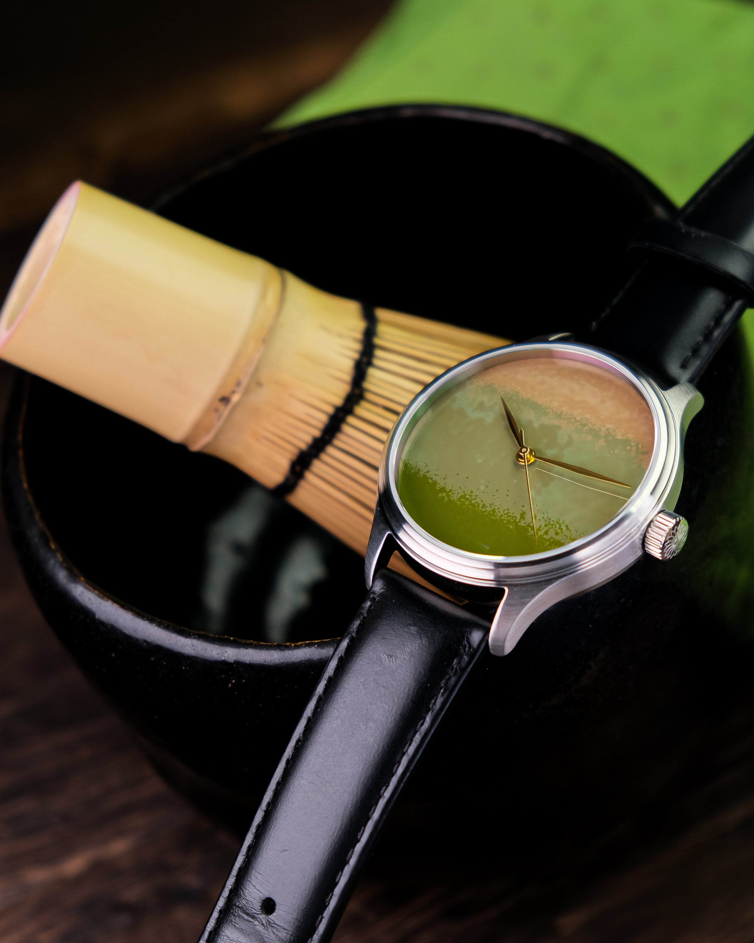 Wristwatch with green leaf design on face next to a bamboo whisk and black container Dream Watch Chawan iro Matcha beside a Matcha Bowl and Matcha Whisk Urushi watches, Urushi Maki-e Urushi dial Wancher Watch 