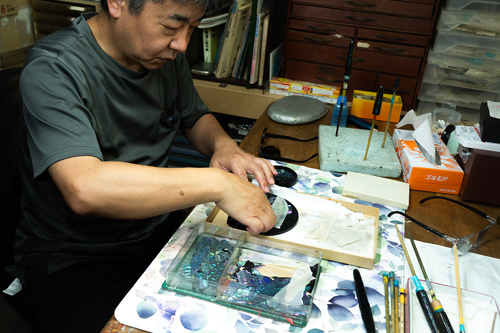 Master Yamanoshita of Taya Shikitten Wancher Watch  wearing a dark gray t-shirt is seated at a cluttered desk, carefully working on cutting shell mother of pearl to create Raden pieces to make urushi raden maki-e watch for Wancher Watch 