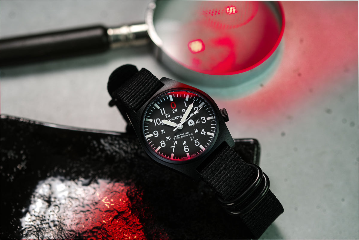 A Wancher Gurkha tilted dial wristwatch with a black fabric strap and a black dial featuring white numerals and red accents, styled on a dark surface with dramatic red lighting and a magnifying glass in the background.