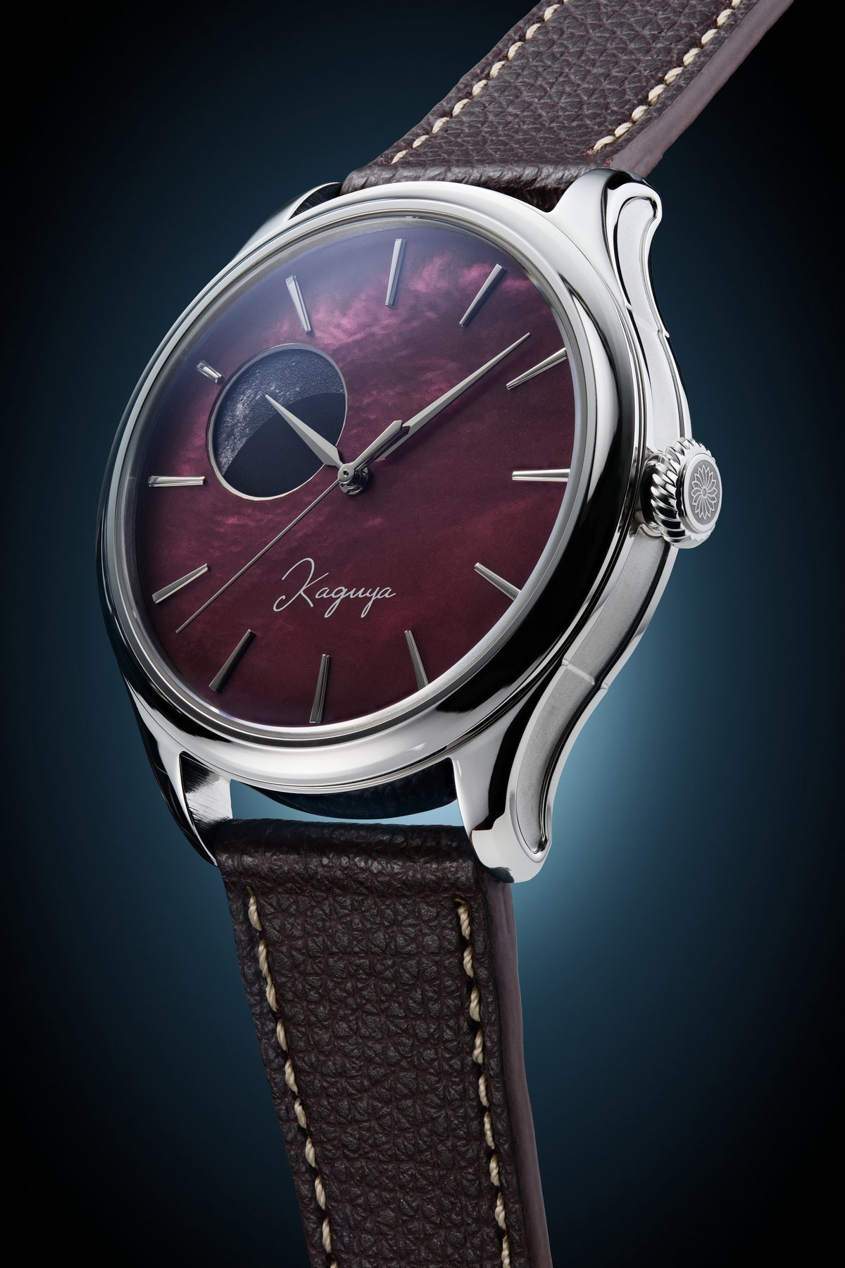 A Dream Moonphase Kaguya AKANE wristwatch with a polished silver-tone case, a deep burgundy dial featuring a moonphase indicator, and a textured brown leather strap with light-colored stitching, set against a dark blue gradient background.