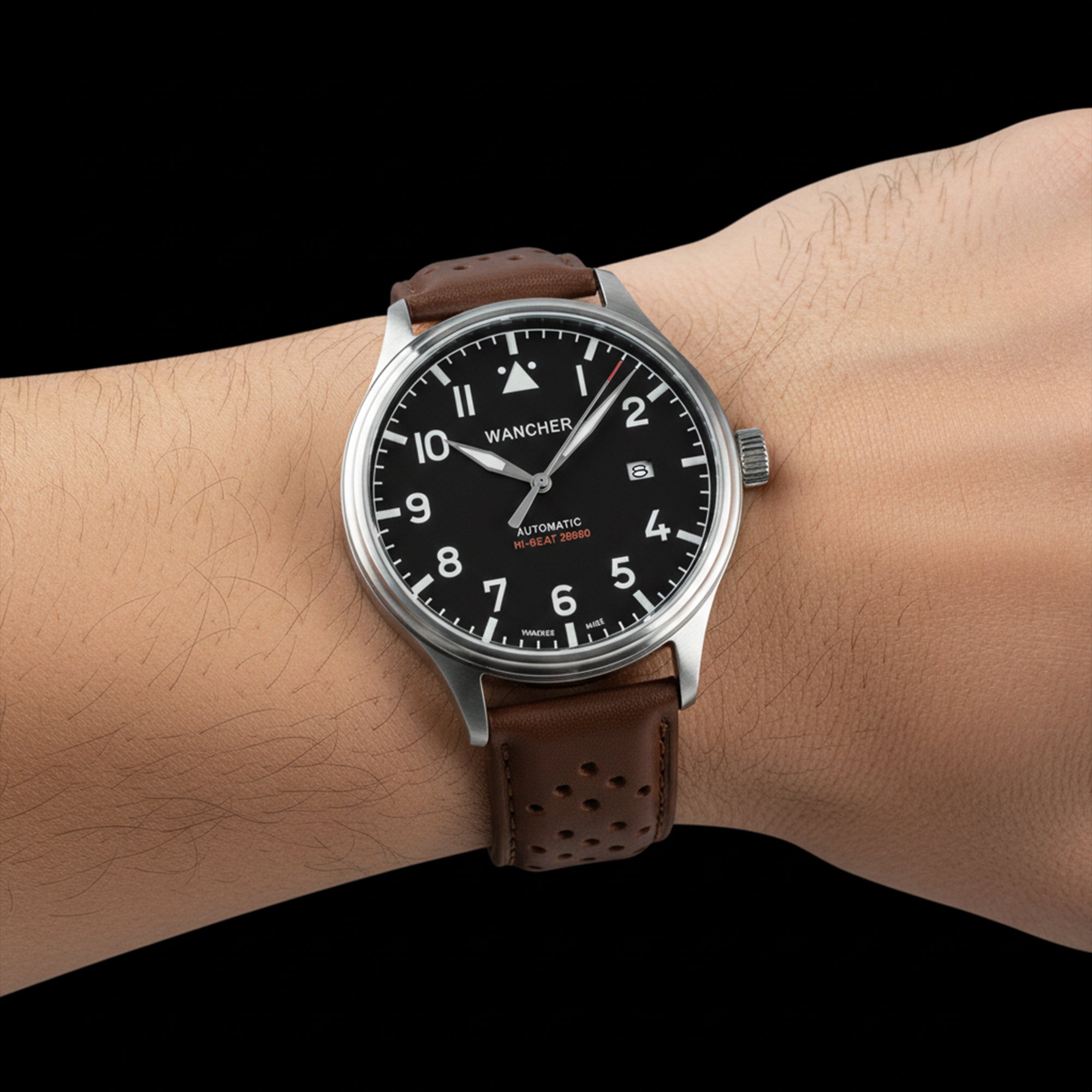A person's wrist wearing the Wancher Flugel Automatic Watch, a pilot-style timepiece with a contrasting black dial and warm brown leather strap.