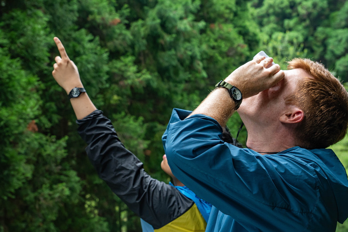 A man in a blue rain jacket and black long-sleeve shirt points upward while holding a small white cup to his mouth, set against a dense green forest background wearing Wancher Gurkha Watch ultimate field watch 