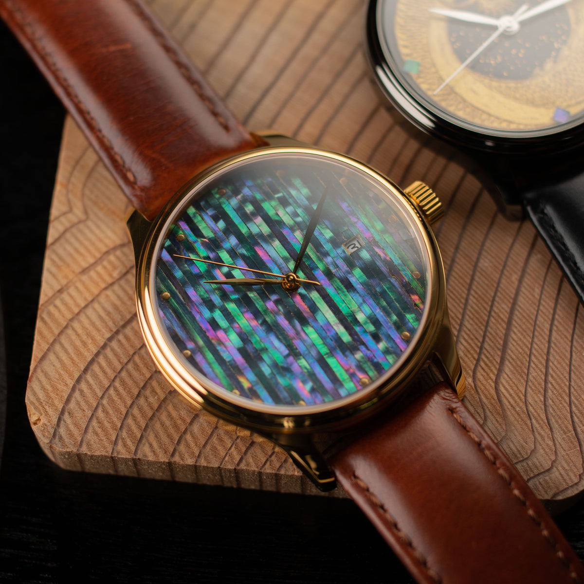 A Dream Watch Raden Pillar - Automatic Urushi maki-e raden watch by wancher watch with a brown leather strap and a gold-tone case, featuring a colorful digital matrix-style dial with green, blue, purple, and pink illuminated elements. The watch is displayed on a wooden block with another watch partially visible in the background.