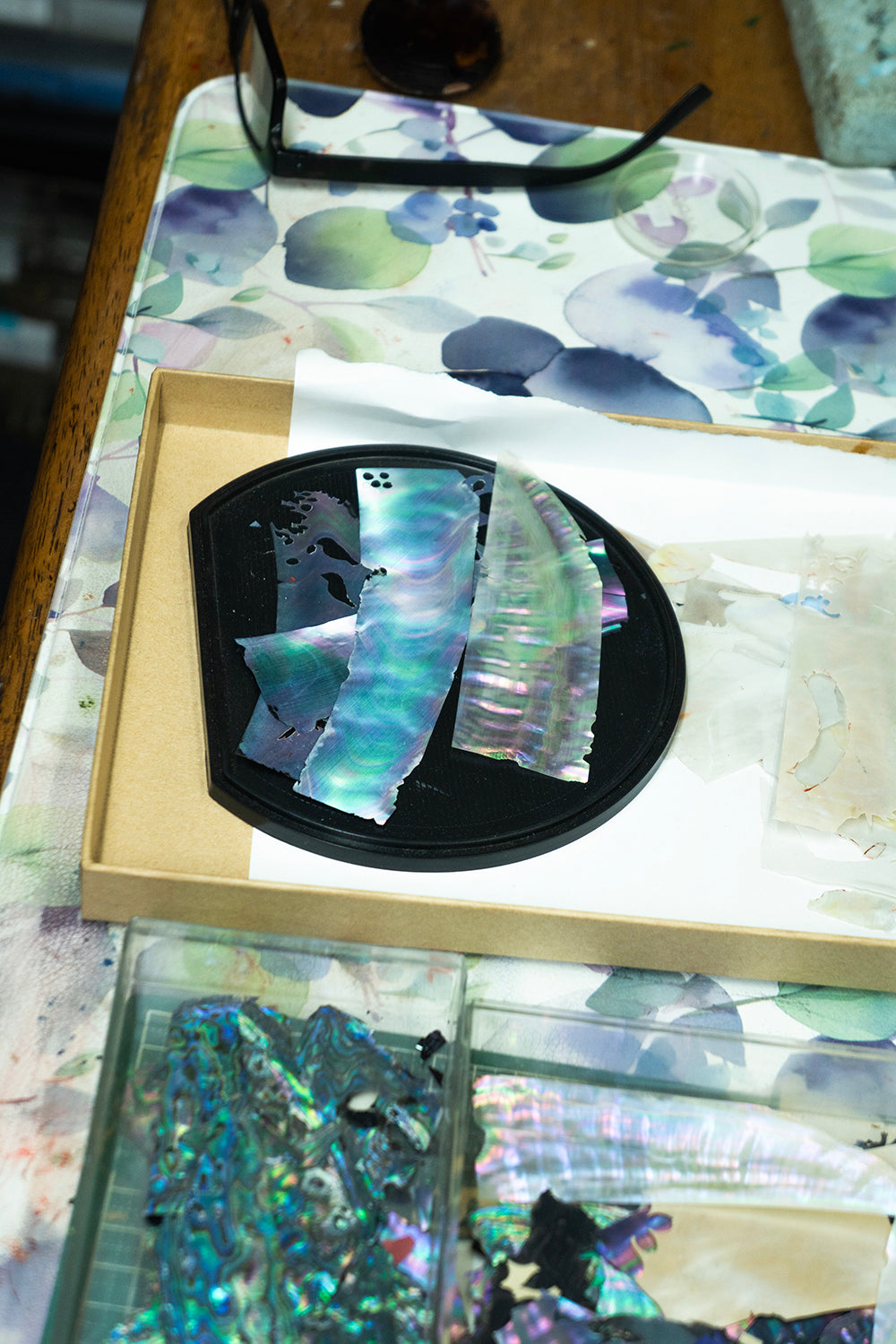 Abalone shell pieces with iridescent blue, green, and purple hues displayed on a black dish inside a tan box, with more shell fragments in clear containers on a floral-patterned surface. Wancher Watch 